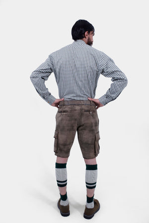 A man with a beard is standing with his back to the camera, hands on hips, wearing a grey and white checkered shirt and grey leather lederhosen. He is also wearing green and grey knee-high socks and brown shoes. The lederhosen has pockets on both sides and is detailed with stitching.