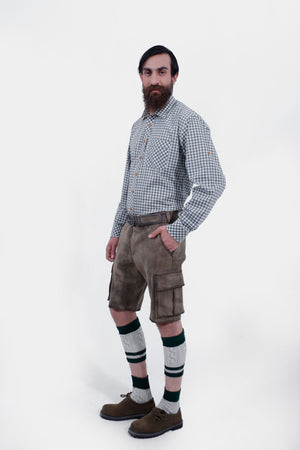 A man with a beard is standing in a three-quarter view against a white background, wearing a grey and white checkered shirt and grey leather lederhosen. He has his right hand in the pocket of the lederhosen, and is also wearing green and grey knee-high socks and brown shoes.