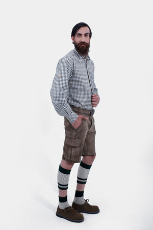A man with a beard is standing in a three-quarter view against a white background, wearing a grey and white checkered shirt and grey leather lederhosen. He has his right hand in the pocket of the lederhosen, and is also wearing green and grey knee-high socks and brown shoes.