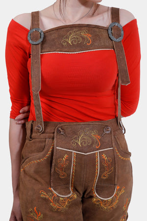 Close-up image of a woman wearing Heidi Klum Lederhosen with suspenders, highlighting the natural-colored embroidery on the bib and legs, paired with a bright red off-the-shoulder top.