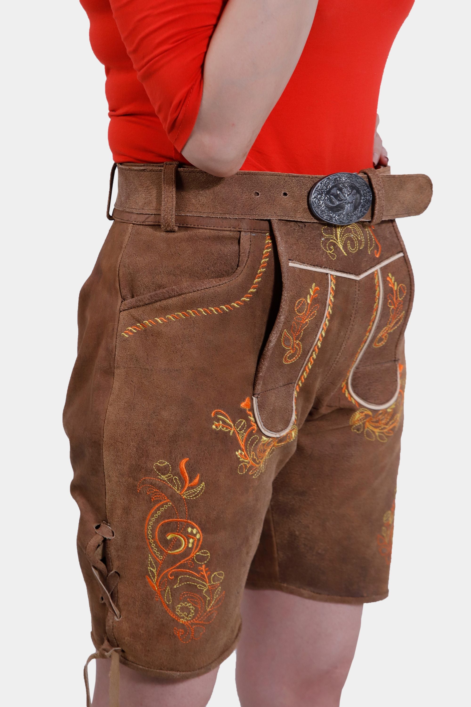 Side view of brown leather Lederhosen shorts showcasing detailed floral embroidery, a brown belt with a silver buckle, and lacing on the side, paired with a bright red top.