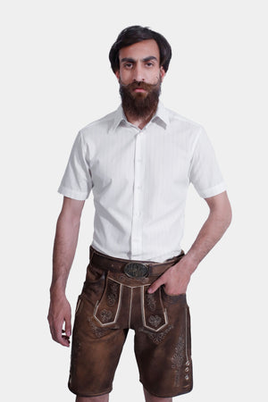 A man poses in a white short-sleeved shirt paired with brown leather lederhosen shorts. He stands with one hand in his pocket.