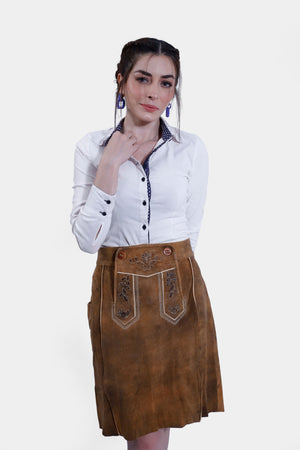 A woman wearing a white shirt with a blue dotted collar and cuffs, paired with a brown leather Jennifer Lederhosen Skirt embroidered with intricate patterns, posing with her hand near her neck.
