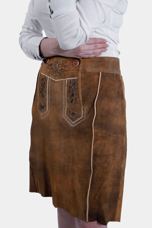 A side view of a woman in a brown leather Jennifer Lederhosen Skirt with detailed embroidery, paired with a white shirt, her arms crossed.
