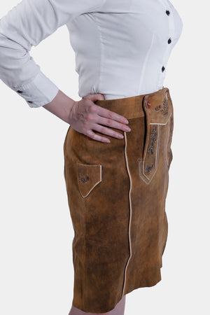 A close-up side view of a woman in a brown leather Jennifer Lederhosen Skirt with detailed embroidery, paired with a white shirt, her arms crossed.