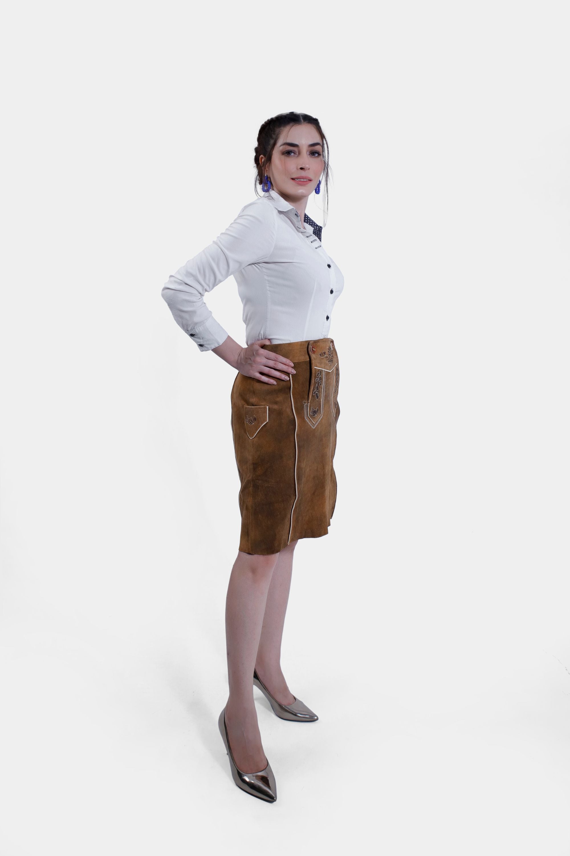 Side profile of a woman wearing a white shirt and a brown leather Jennifer Lederhosen Skirt with intricate embroidery, standing with one arm crossed and her hand near her chin.
