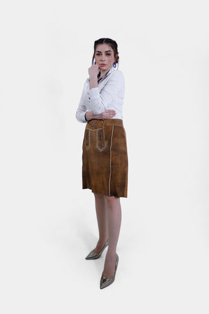 A woman wearing a white shirt and a brown leather Jennifer Lederhosen Skirt with intricate embroidery, standing with one arm crossed and her hand near her chin.