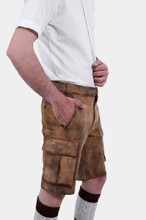 A man with a beard is standing in a three-quarter view against a white background, wearing a short-sleeved white shirt and brown leather lederhosen. He has his right hand in the pocket of the lederhosen, and is also wearing brown and grey knee-high socks and brown shoes.