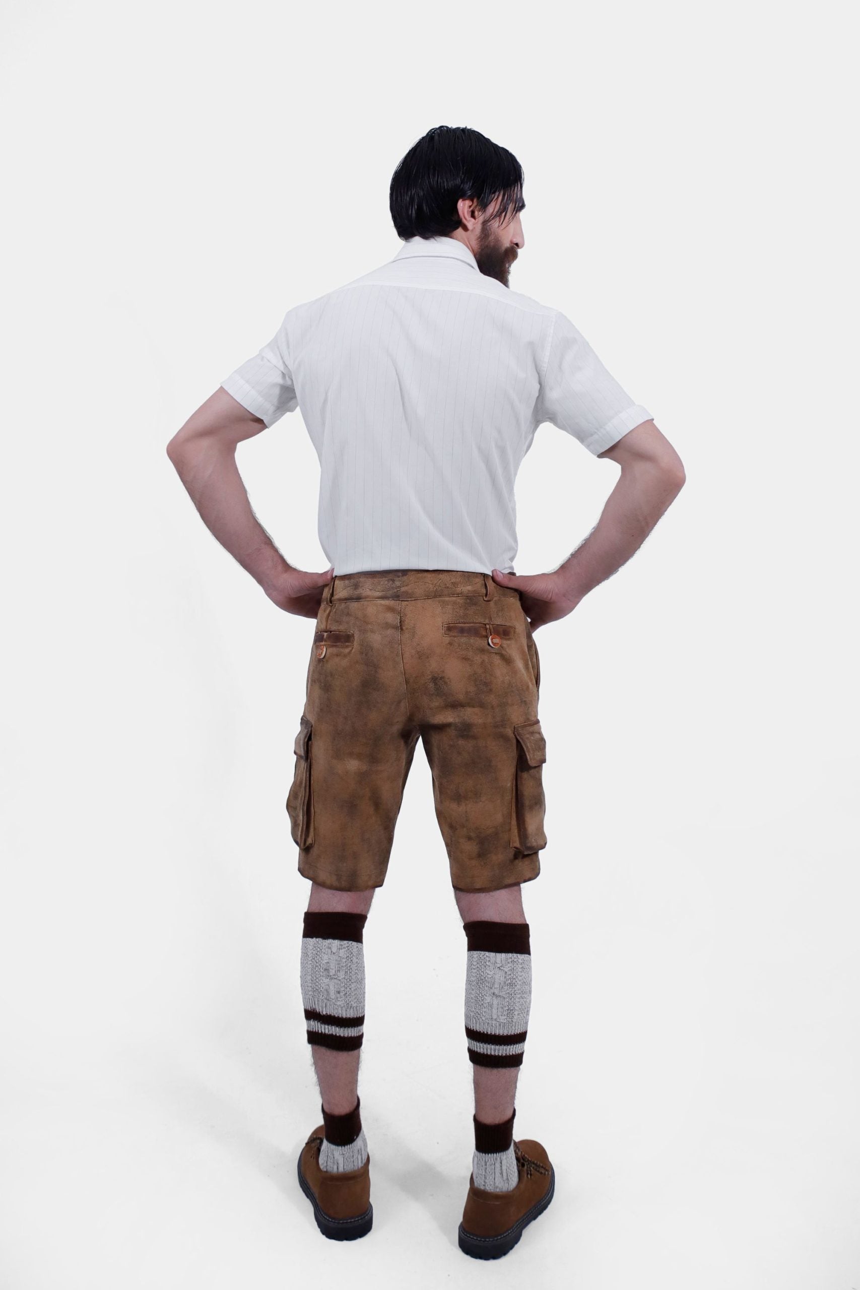 A man with a beard is standing with his back to the camera, hands on hips, wearing a short-sleeved white shirt and brown leather lederhosen. He is also wearing brown and grey knee-high socks and brown shoes. The lederhosen has pockets on both sides and is detailed with stitching.