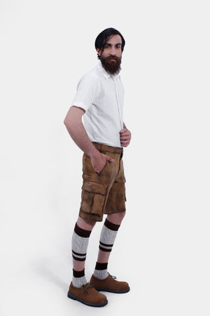 side view of a man wearing brown leather cargo lederhosen and a short-sleeved white shirt. The man has his right hand in the pocket, highlighting the detailed stitching and buttons on the lederhosen.