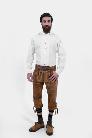 A man with a beard is wearing traditional Bavarian Bundhosen. He is dressed in a white shirt with brown leather knee-length trousers adorned with intricate embroidery. The outfit is completed with traditional Bavarian shoes and socks.