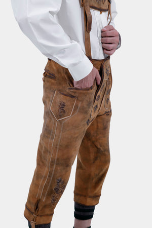 A side view of a man wearing traditional brown leather Bundhosen with intricate embroidery. The Bundhosen are paired with a white shirt and the outfit is accessorized with a leather belt and traditional shoes.