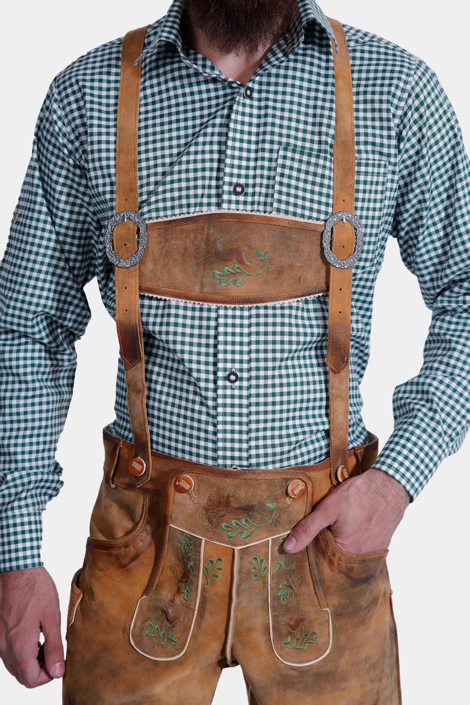 A man wearing a Lars Eidinger Lederhosen outfit, including tan leather shorts with green embroidery, a green checkered shirt, and traditional leather suspenders, standing with hands in pockets.