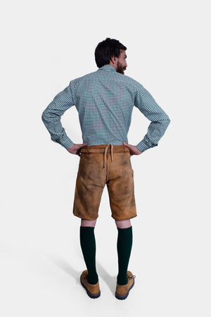 Back view of a man in a Lars Eidinger Lederhosen outfit, showing the rear details of the tan leather shorts and the fit of the green checkered shirt.