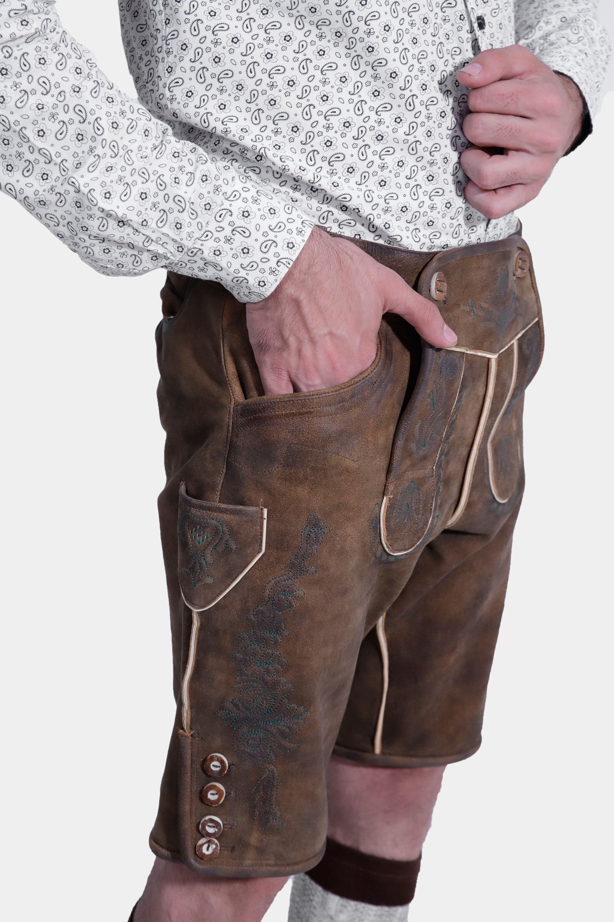 Closed side view of a man wearing traditional Bavarian Lederhosen. The outfit includes a white shirt with a subtle pattern and brown leather shorts with intricate embroidery. He completes the look with knee-high woolen socks and brown shoes. The attire captures the classic Bavarian style.