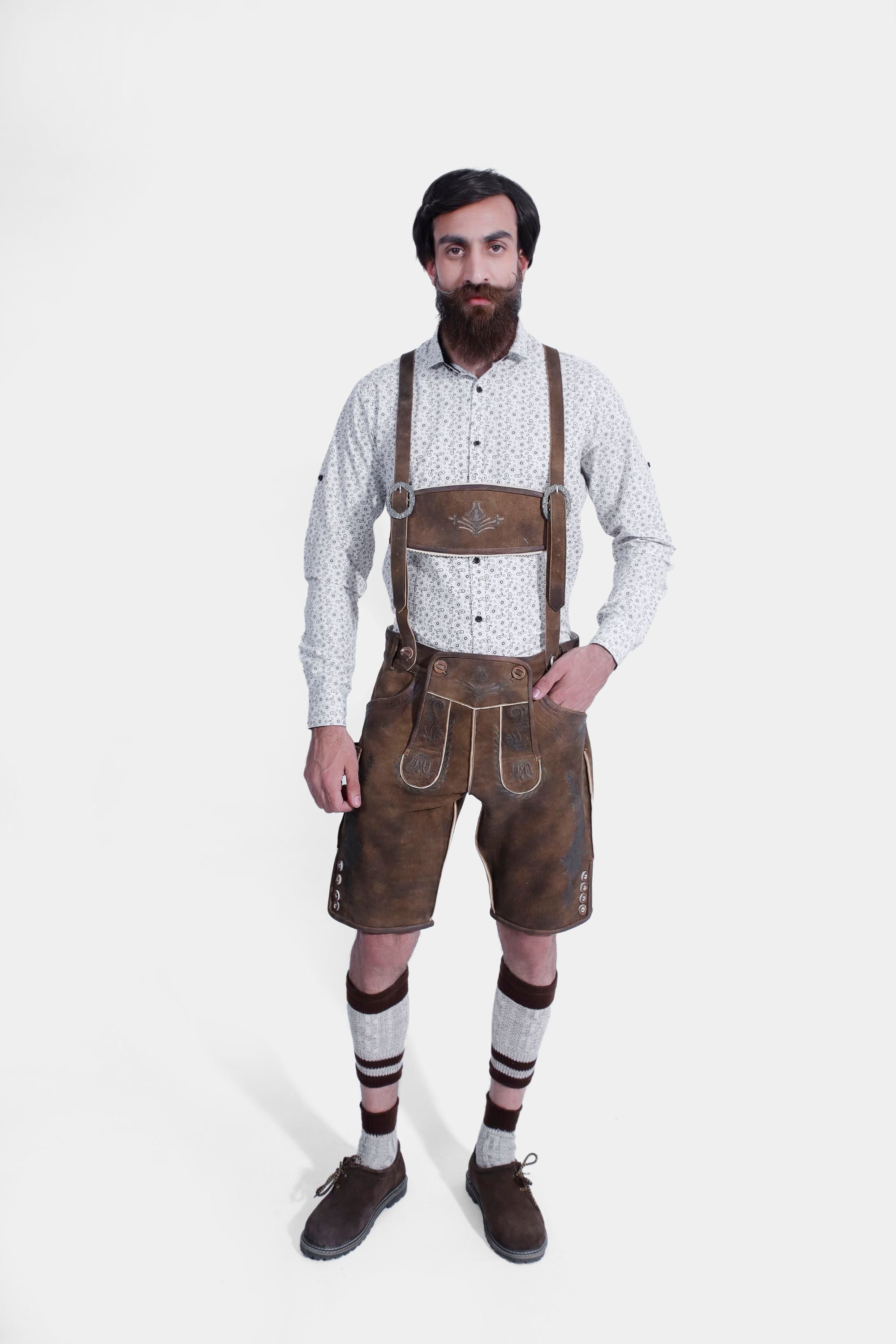 A man wearing traditional Bavarian Lederhosen. The outfit includes a white shirt with a subtle pattern, brown leather shorts with intricate embroidery, and suspenders. He completes the look with knee-high woolen socks and brown shoes. The attire captures the classic Bavarian style.