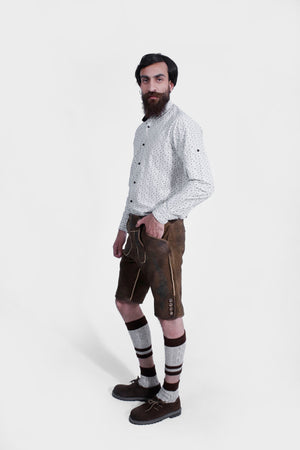 Side view of a man wearing traditional Bavarian Lederhosen. The outfit includes a white shirt with a subtle pattern and brown leather shorts with intricate embroidery. He completes the look with knee-high woolen socks and brown shoes. The attire captures the classic Bavarian style.