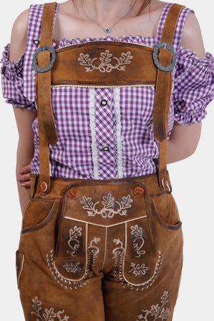 Close-up of a model wearing brown leather lederhosen with intricate white embroidery on the front, paired with a purple checkered blouse.