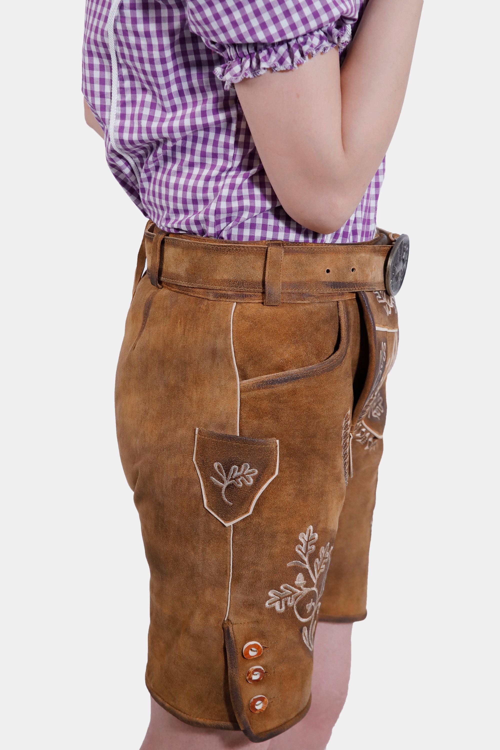 Side view of a model in brown leather lederhosen shorts with white embroidery, showcasing the side pocket and button detailing, paired with a purple checkered blouse