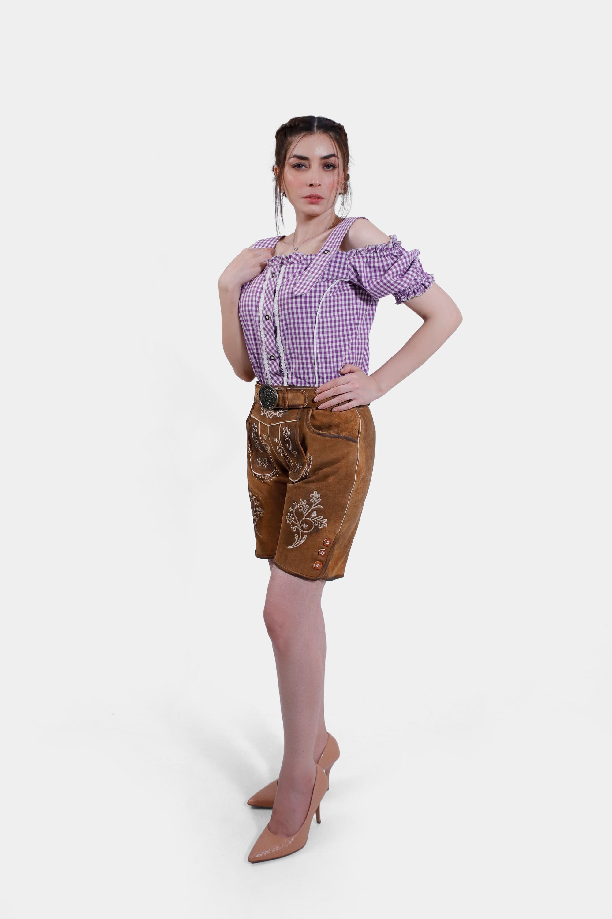 Model striking a pose in brown leather lederhosen shorts with white embroidery, paired with a purple checkered blouse and nude high heels, seen from the side.