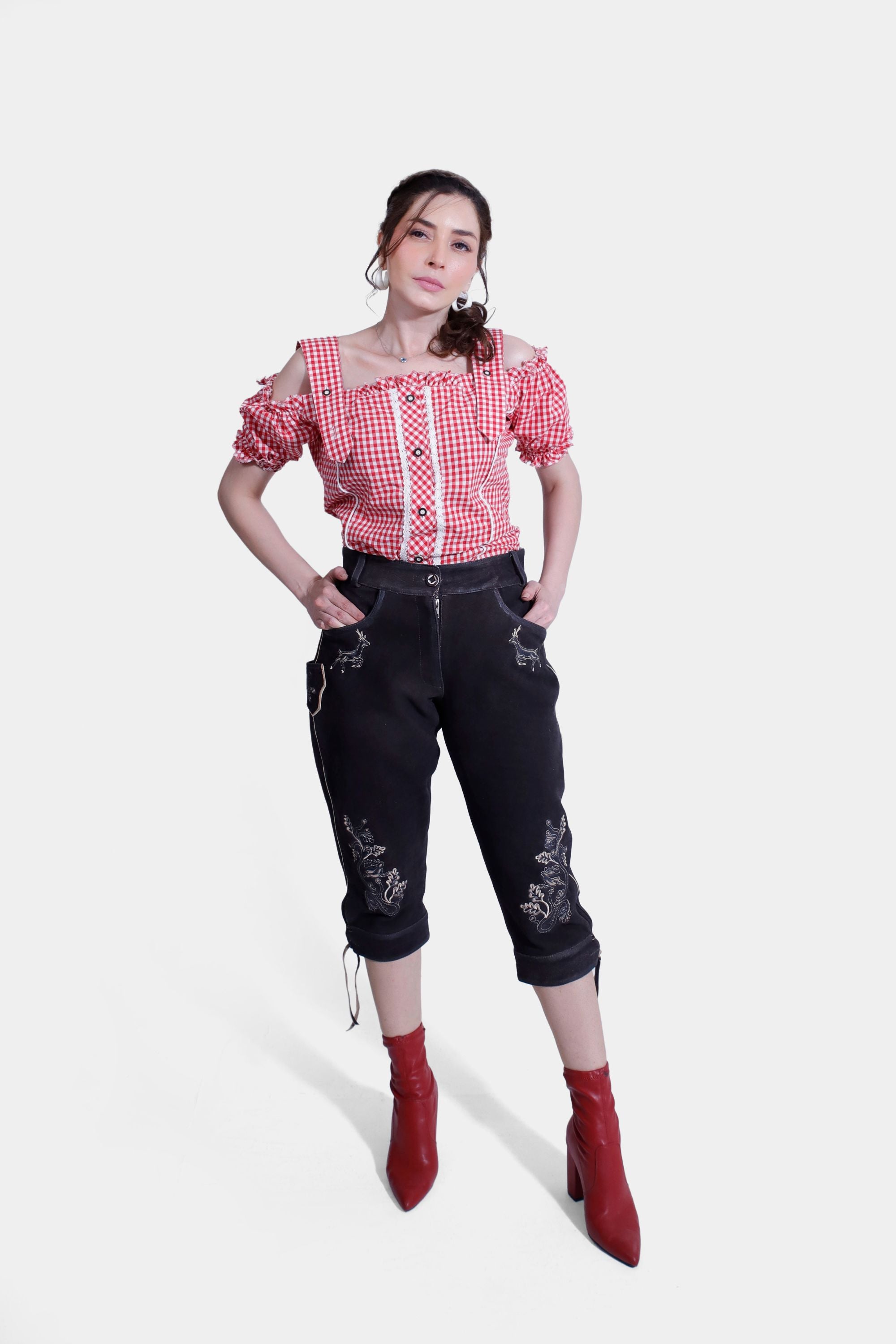 The woman strikes a full-body pose, showcasing her outfit that includes a red and white checkered off-shoulder blouse and black knee-length lederhosen pants with detailed embroidery. She is wearing red high-heeled ankle boots and has her hands casually placed in her pockets.