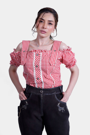 A closer shot of the woman in a red and white checkered off-shoulder top and black lederhosen pants, with intricate embroidery visible on the pants. Her expression is calm, and her hands rest inside her pockets.