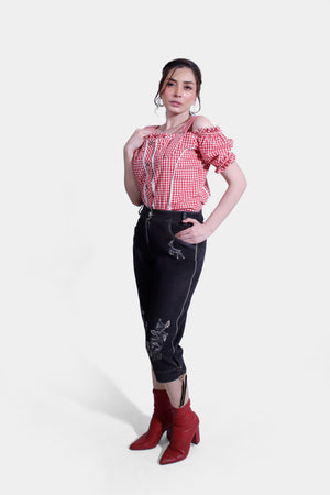 side view of woman wearing a red and white checkered off-shoulder blouse with ruffled sleeves and black embroidered lederhosen pants, standing with hands in pockets.
