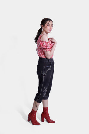 Another side view of woman wearing a red and white checkered off-shoulder blouse with ruffled sleeves and black embroidered lederhosen pants, standing with hands in pockets.