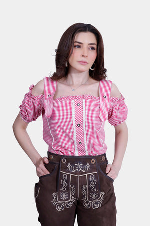 A woman is wearing a pink and white checkered off-shoulder blouse with button details, paired with brown embroidered lederhosen shorts. Her hands are casually placed in the pockets, and her hair is styled in loose waves. The lederhosen feature intricate floral embroidery on the front.