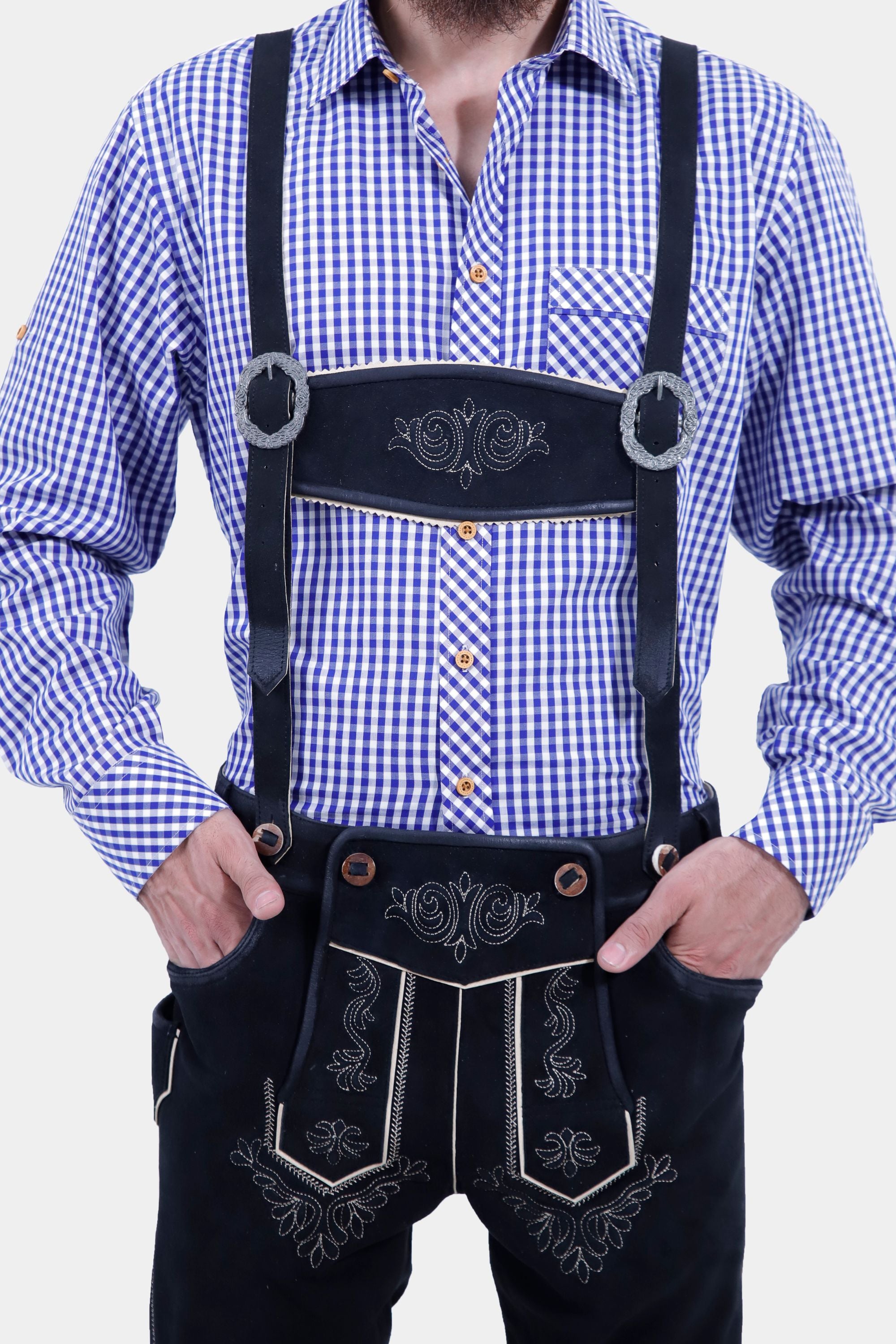 Close-up view of a man wearing a blue and white checkered shirt, showcasing the detailed buttons and front pocket. He is also wearing black leather bundhosen with suspenders and intricate embroidery, with his hands resting on his hips.