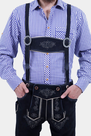 Close-up view of a man wearing a blue and white checkered shirt, showcasing the detailed buttons and front pocket. He is also wearing black leather bundhosen with suspenders and intricate embroidery, with his hands resting on his hips.