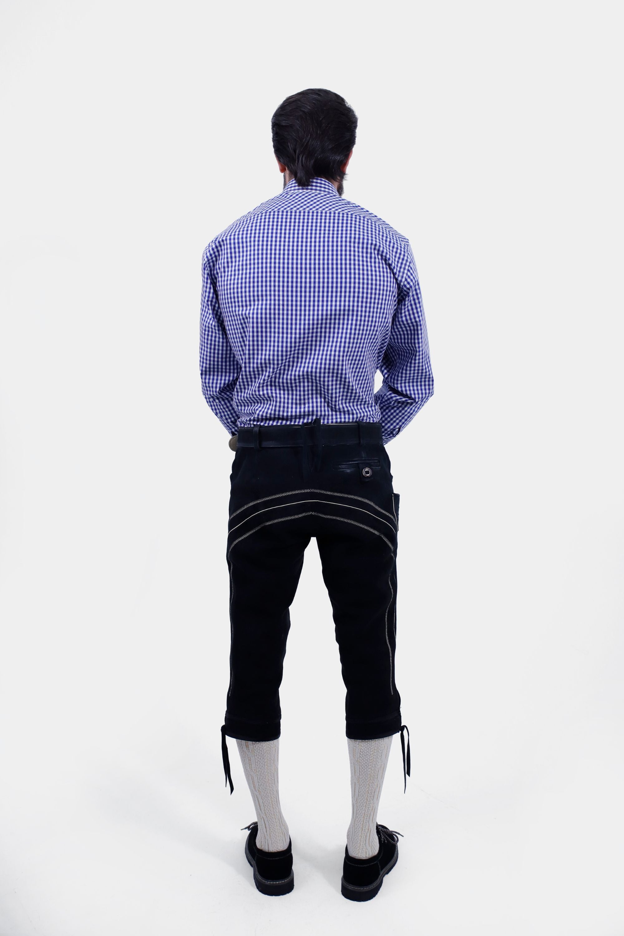 Rear view of a man wearing a blue and white checkered shirt and black leather bundhosen, showing the back pockets and overall fit of the bundhosen along with cream-colored knee-high socks.