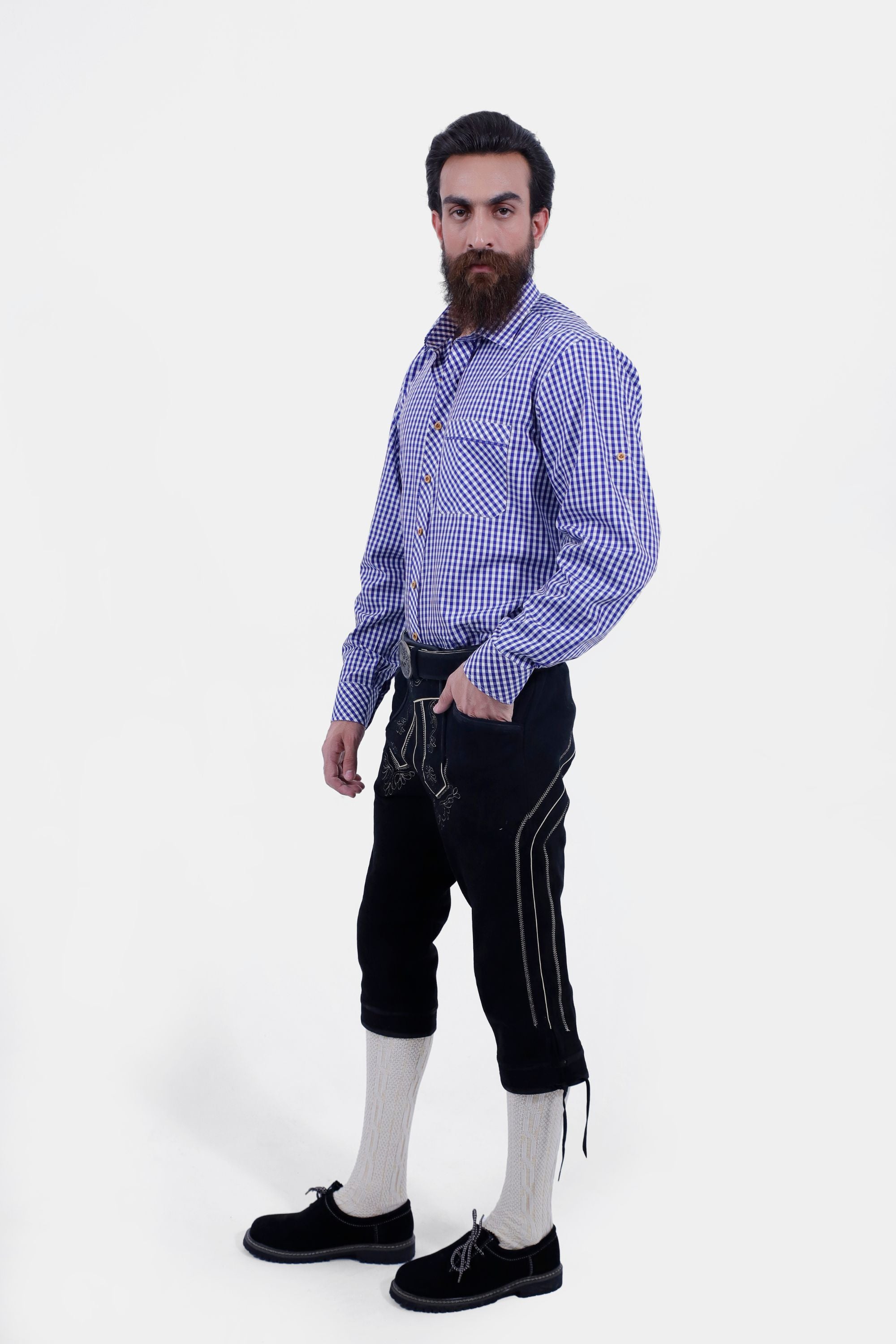 A man with a beard and styled hair wearing a blue and white checkered shirt and black leather bundhosen, standing with a slight side angle and his hand in his pocket, showcasing the bundhosen's detailed craftsmanship and cream-colored knee-high socks.