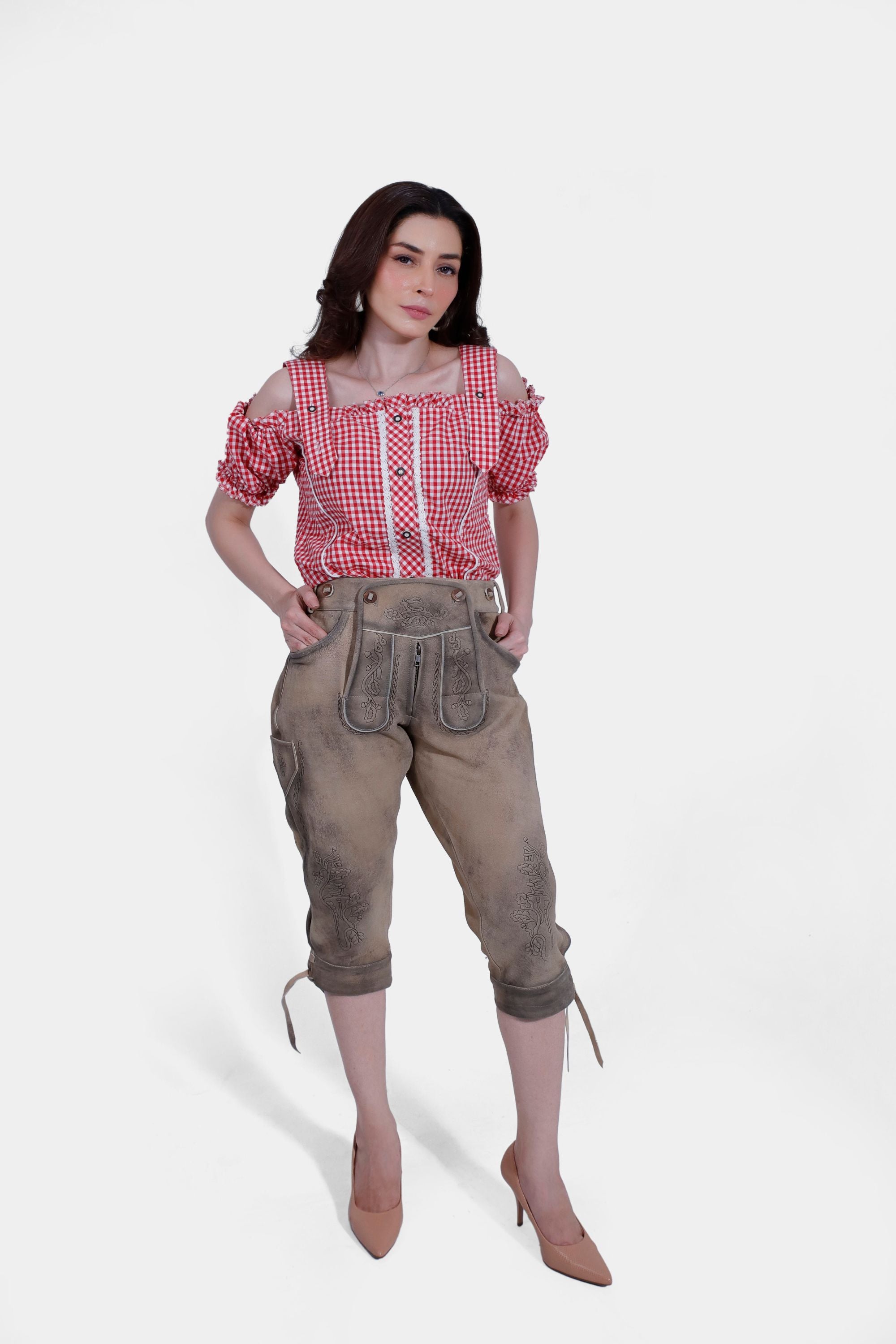 A woman wearing a red and white checkered off-shoulder blouse with ruffled sleeves and light brown lederhosen pants, standing with hands in pockets.