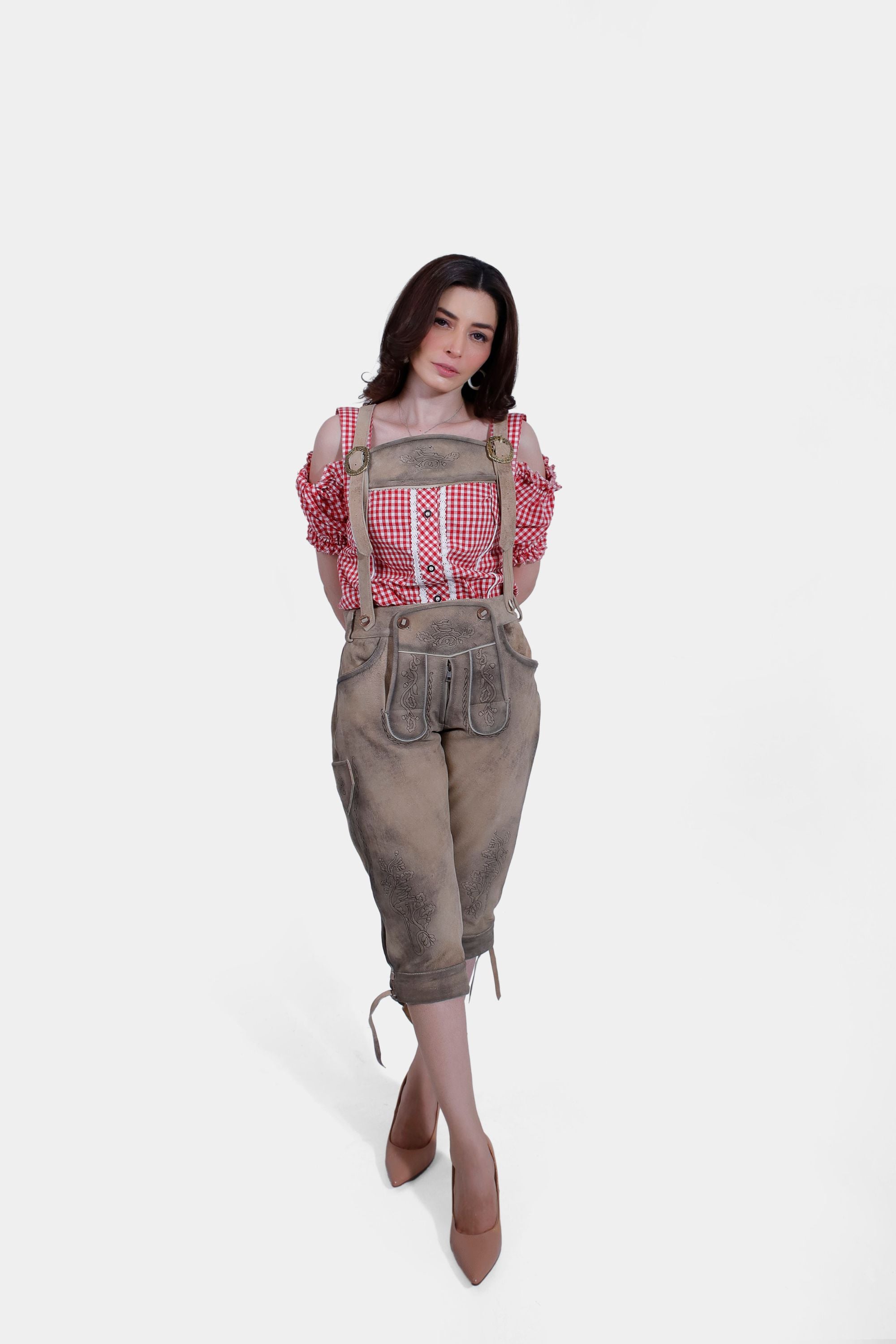 Woman in a red and white checkered off-shoulder blouse and light brown embroidered lederhosen pants, standing with hands behind her back.