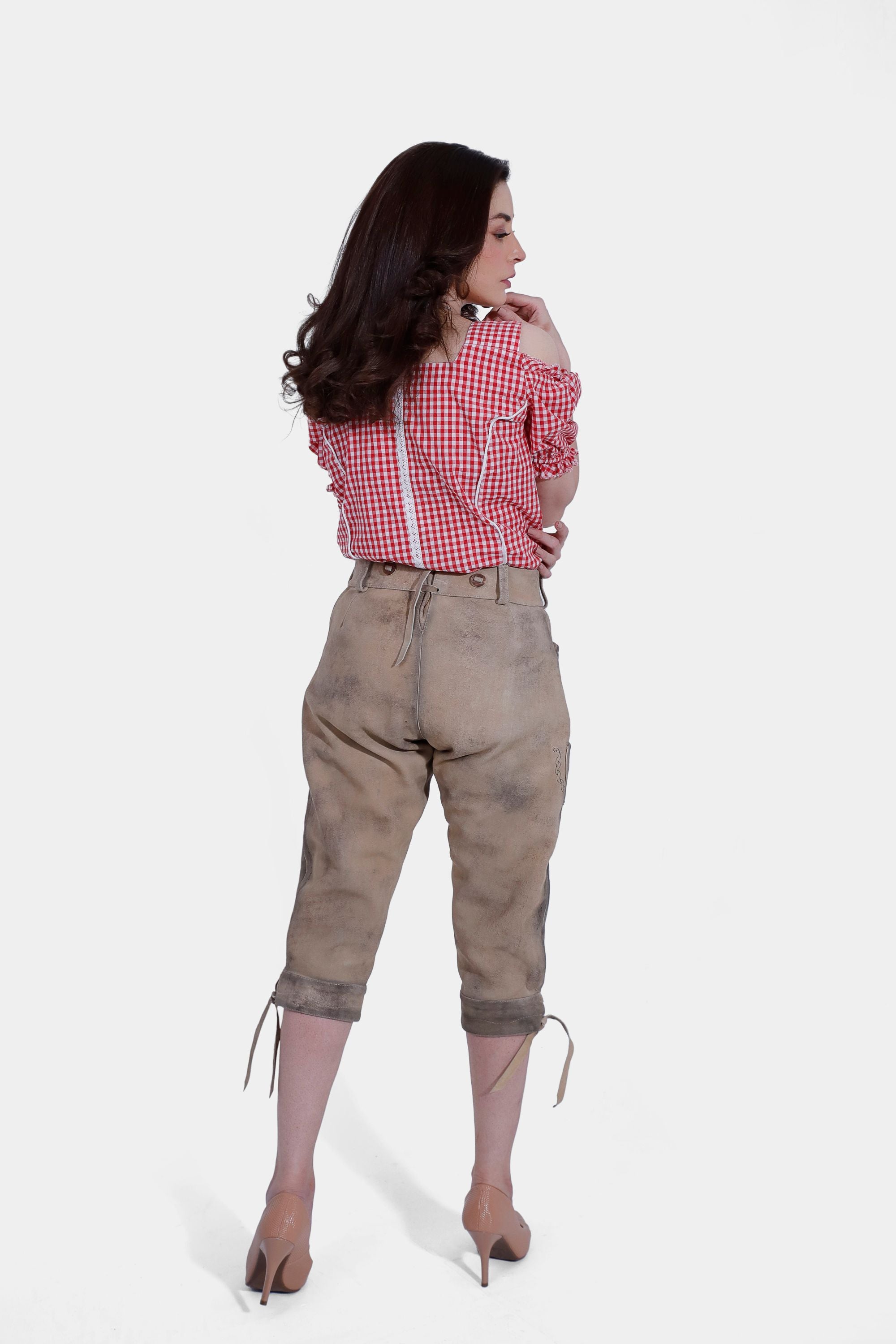 back view of woman in a red and white checkered off-shoulder blouse and light brown embroidered lederhosen pants, standing with hands behind her back.