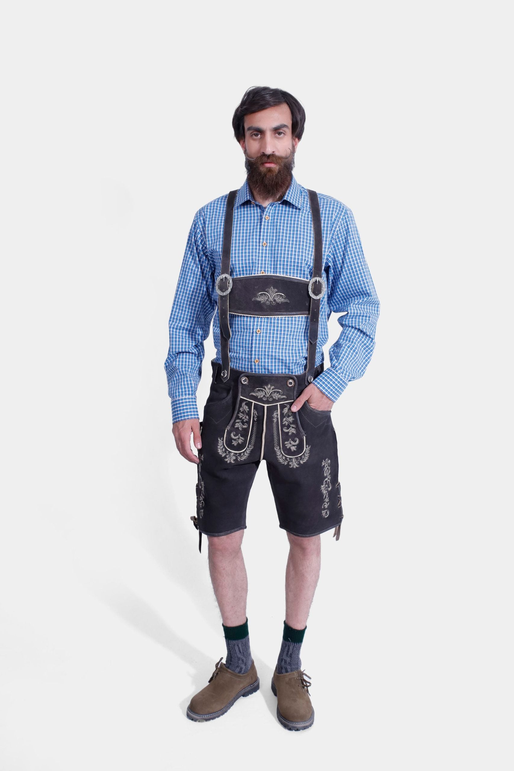 A man wearing traditional Bavarian Lederhosen. He is dressed in a blue checkered long-sleeve shirt paired with black leather shorts featuring intricate white embroidery and suspenders. The look is completed with dark green knee-high socks and brown shoes, embodying a classic Bavarian style.