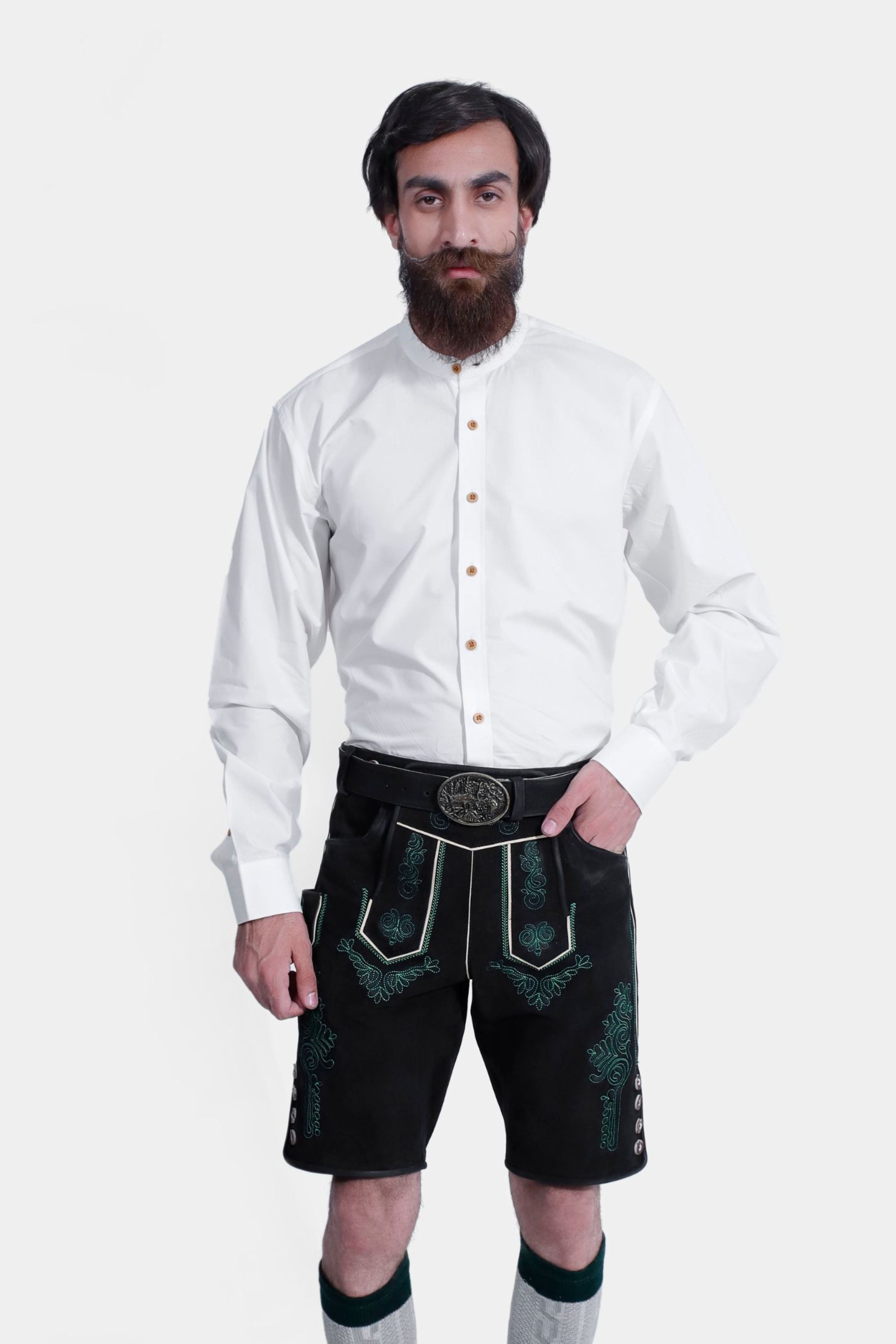 Full-body view of the same man wearing the white shirt and black lederhosen with green embroidery. He also has gray and green knee-high socks and black shoes, standing against a plain white background.