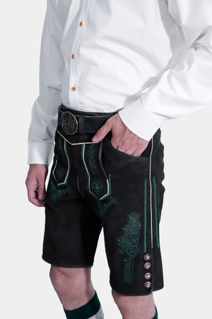 A side view close-up of the man's hand resting in the pocket of the black lederhosen, highlighting the green embroidery and side buttons. He is wearing a white button-up shirt, and the background is plain white.