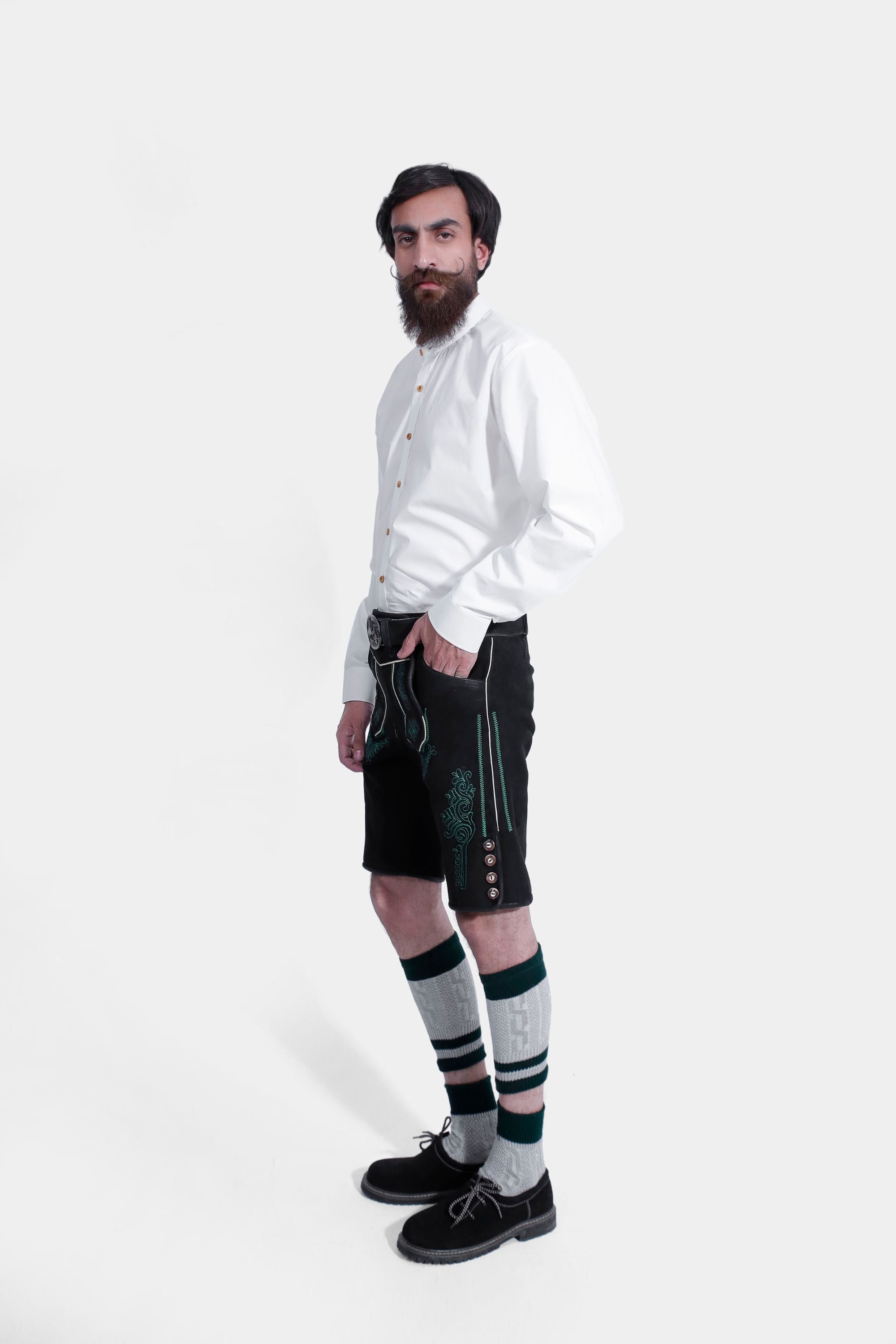 Side view of a man in a white shirt and black lederhosen with green embroidery, one hand in pocket, against a white background.