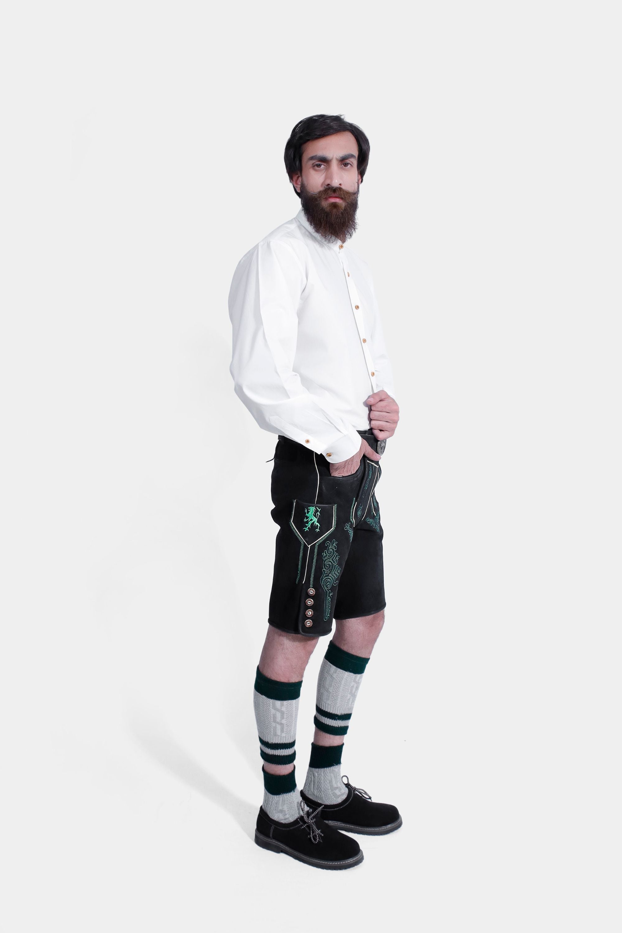 Side view of a man in a white shirt and black lederhosen with green embroidery, one hand in pocket, against a white background.