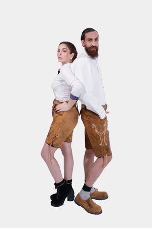The couple standing back-to-back in their Neuschwanstein Couple Outfits. The woman wears a white blouse with golden-brown leather shorts that feature detailed embroidery, and black lace-up boots. The man is in a matching white button-down shirt and golden-brown leather shorts with intricate embroidery, paired with brown shoes and green knee-high socks.