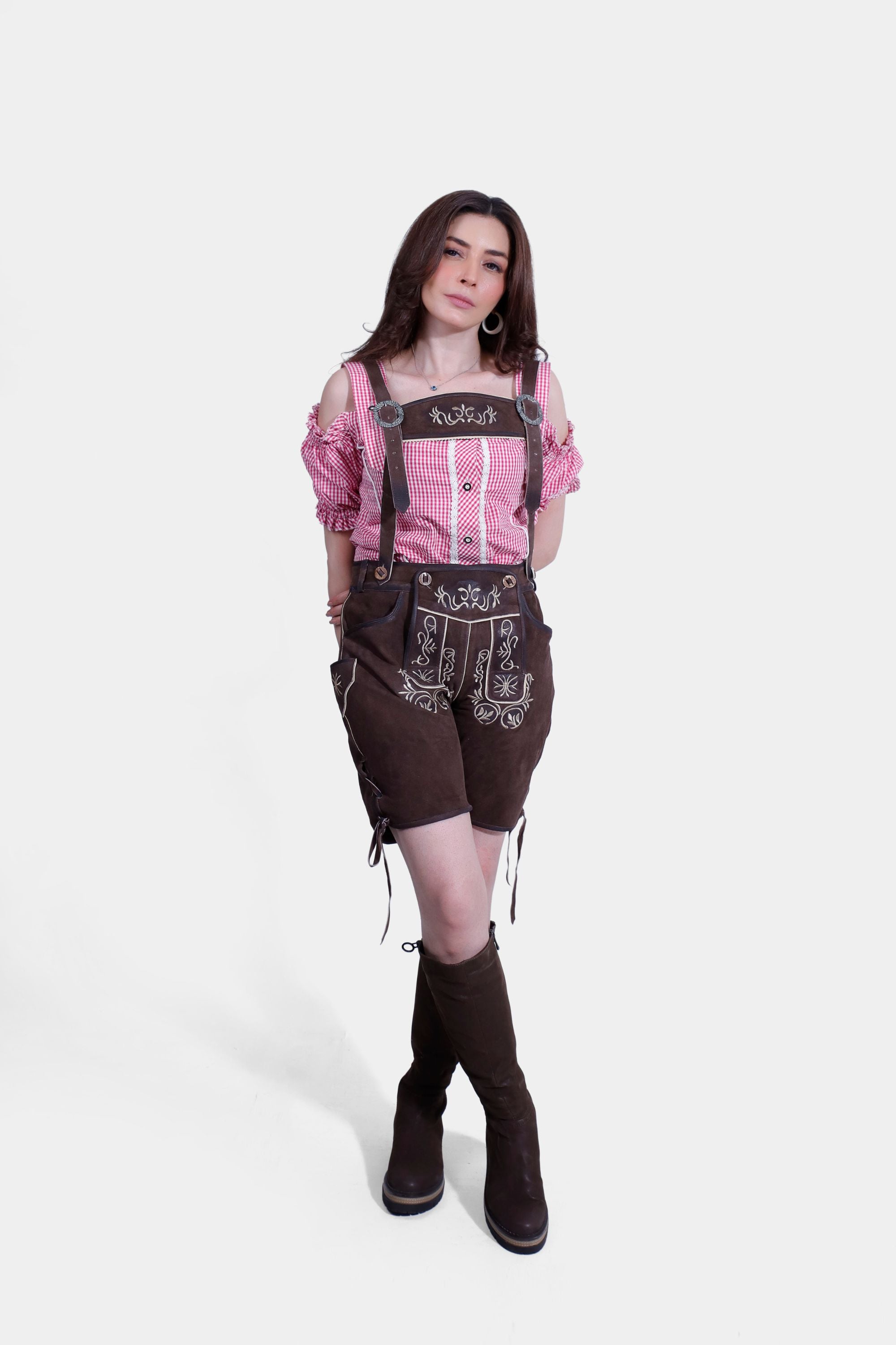 A woman wearing a red and white checkered off-shoulder blouse with ruffled sleeves and dark brown embroidered leather shorts, standing with her legs crossed and hands on hips.