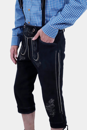 Another side view of the man wearing the blue checkered shirt and black Bundhosen. The detailed embroidery on the Bundhosen is highlighted, and the man has his hand in his pocket.