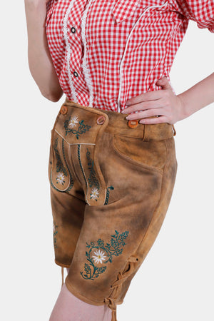 Close-up of a woman wearing a red and white checkered off-the-shoulder top and brown lederhosen with green floral embroidery. The focus is on the lederhosen's intricate embroidery and the woman's hand resting on her hip.