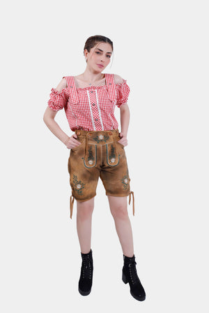 A woman with braided hair wearing a red and white checkered off-the-shoulder top and brown lederhosen with green floral embroidery. She stands facing forward with her hands on her hips, posing confidently.