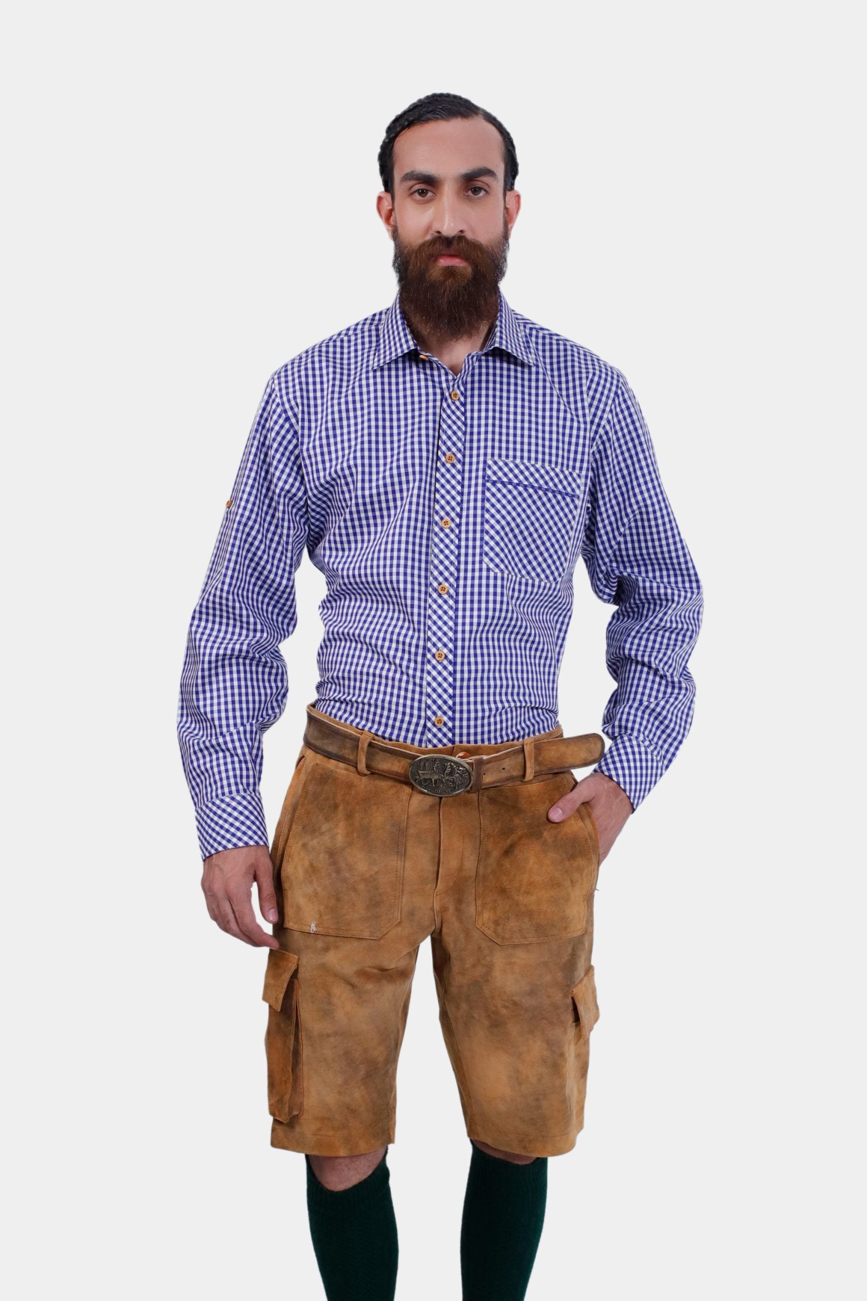 A man with a beard wearing a blue and white checkered dress shirt and traditional brown cargo lederhosen with pockets. He stands facing forward with his left hand in his pocket.