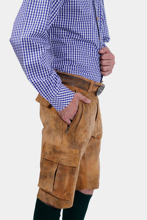 Side profile of a man wearing a blue and white checkered dress shirt and brown cargo lederhosen with pockets. He has his left hand in his pocket and is looking slightly towards the camera.