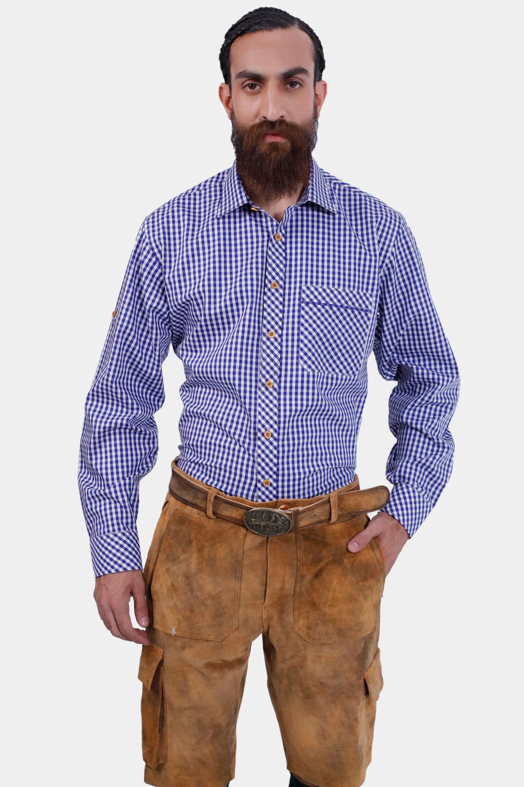 A man with a beard in a blue and white checkered dress shirt and brown cargo lederhosen. He is standing facing the camera with a neutral expression, his left hand in his pocket.