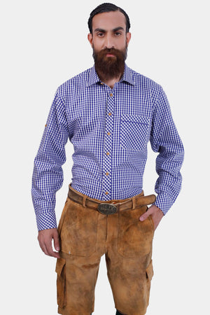 A man with a beard in a blue and white checkered dress shirt and brown cargo lederhosen. He is standing facing the camera with a neutral expression, his left hand in his pocket.